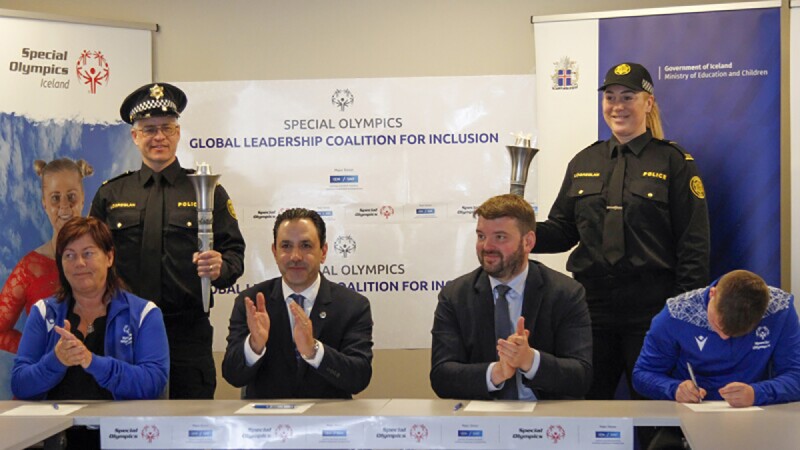 A woman (claps) and three men (two clapping and one signing a document) sit at a table bearing the Special Olympics and SNF logos.