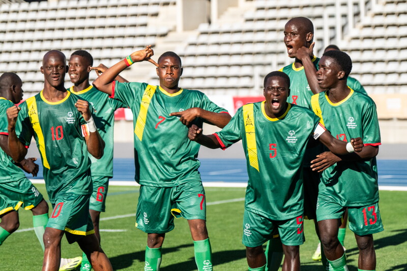 A group of footballers in green uniforms joyfully celebrating on a soccer field, raising their arms and smiling after a victorious goal.