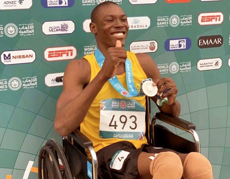 young man in a yellow tank top and black shorts sitting in a wheel chair showing off the medal around his neck.