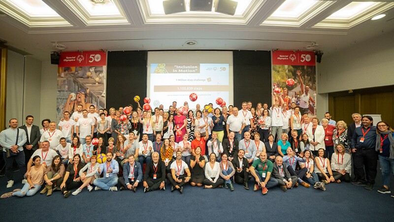 90 participants in a group photo. 4 rows of people are in front of a projection screen with text that reads, inclusion in Motion.