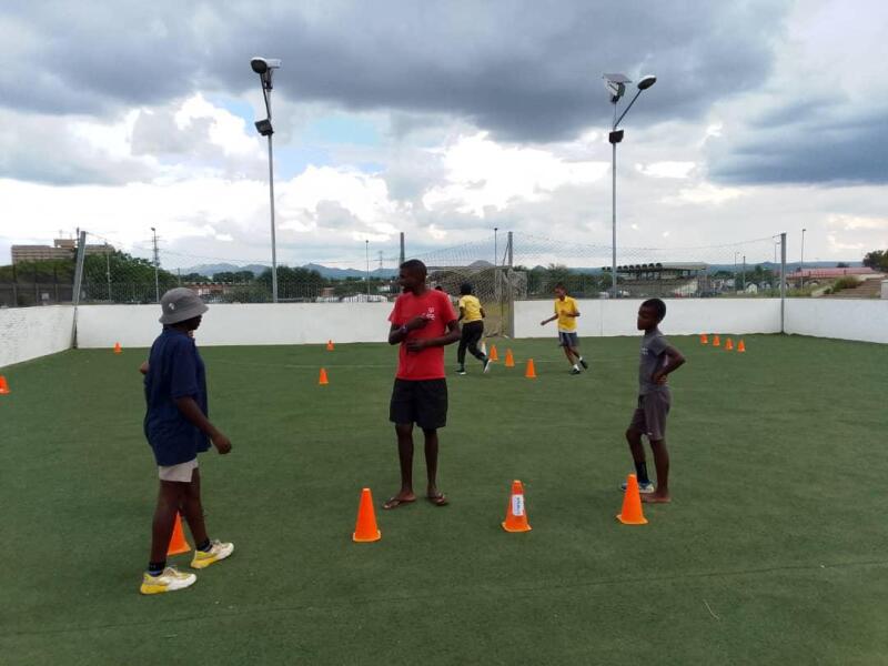 Three individuals standing on an outdoor grass field with orange cones lined up before a fitness drill.