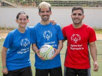Two men, one wearing a red shirt and another a blue one, and a woman wearing a blue shirt pose for the camera while the man in the middle holds a football