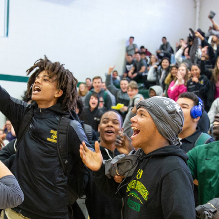 An image of an energetic school assembly. A young man in the front is reaching for a baton while other young adults cheer.