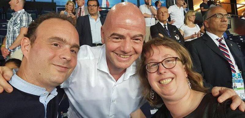 Two men and a women face the camera smiling with a crowd of people in the background. The man in the centre has his arms around the man and woman at either side.