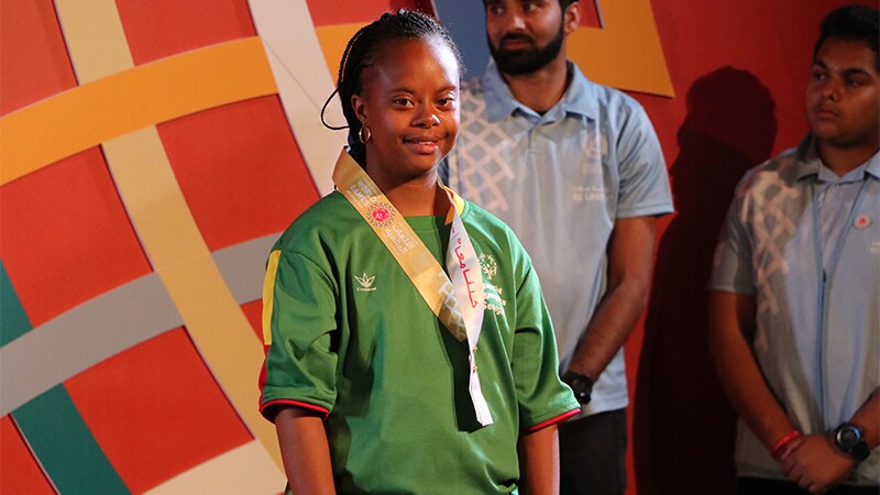 A smile from Team Senegal athlete Khadija Sy after the medal ceremony for the 100m run. She's standing on stage in front of the World Games logo with volunteers behind her.
