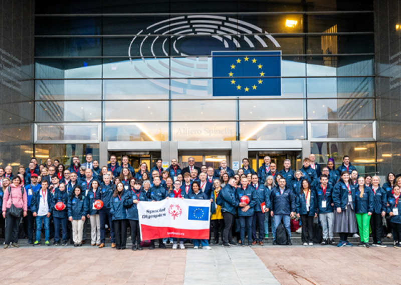 A large group of people stand together with a Special Olympics and European Union branded banner outside a large building.
