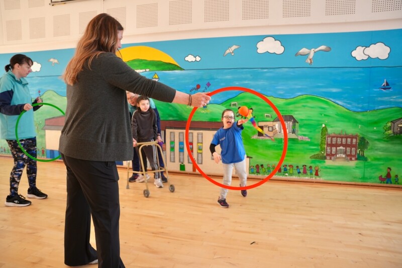 In the foreground, a woman stands holding a red hoop while a child throws a orange ball with a multicolor tail through it. Another child using a mobility walker is nearby. The room has a wooden floor and a brightly painted mural on the wall depicting hills, buildings, clouds, birds, and water.