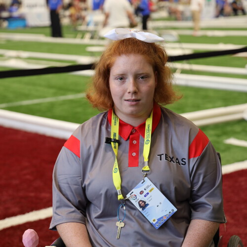 Emileigh Marsh sits in a wheelchair on an indoor turf competition field, wearing a gray and red Texas team shirt and a yellow credential lanyard. Bocce courts and event participants are visible in the background.