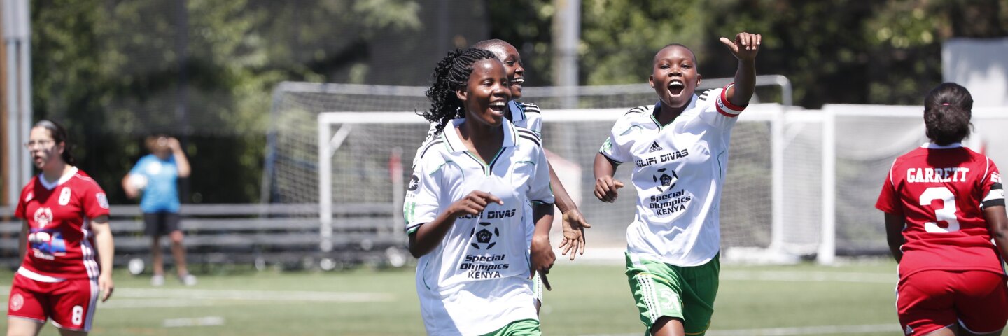 Three girls in white and green on the pitch celebrating and two girls in red.
