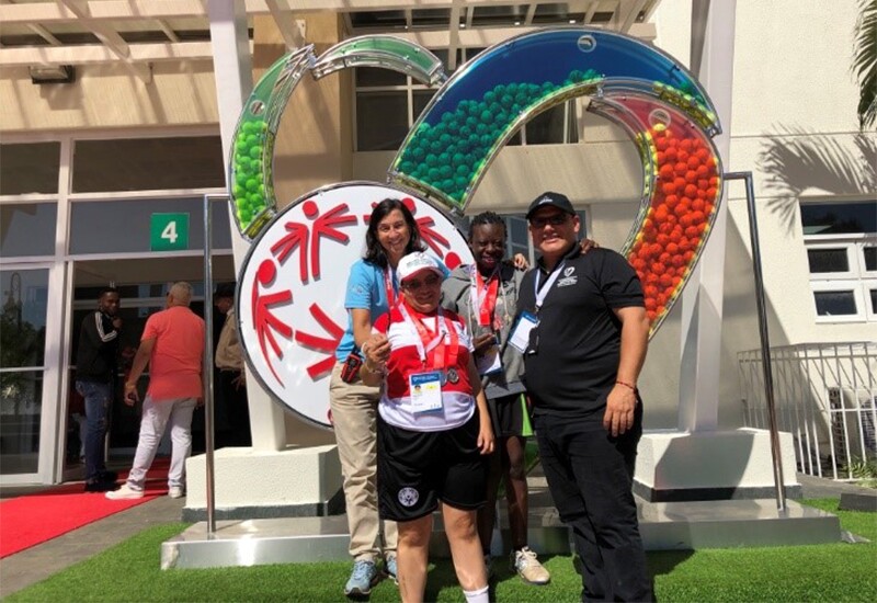 Coach Maria standing in front of the Special Olympics World Tennis Invitational logo installation with two athletes and another coach in front of a pavilion.
