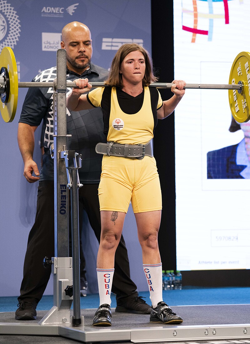 Female power lifter from Cuba in yellow and black holding a bar on her shoulders, a spotter is behind her.