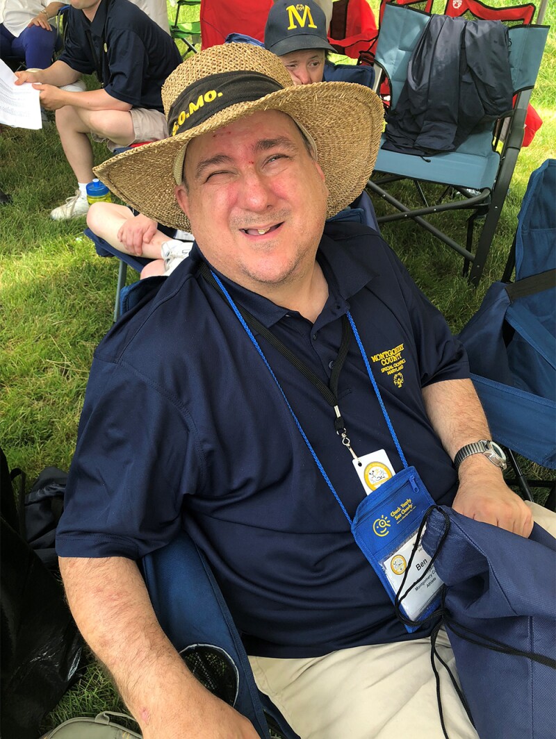 Ben at bocce practice for Special Olympics Maryland. He's sitting down in a folding chair in a blue Montgomery County Special Olympics blue polo, tan pants and straw hat.