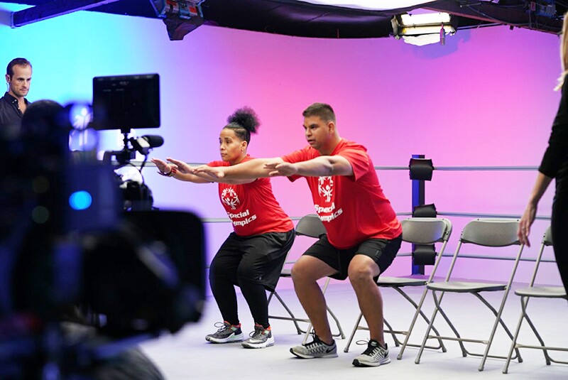 Vince and Angel performing squats in front of a chair; cast, crew, and equipment for filming can be seen.