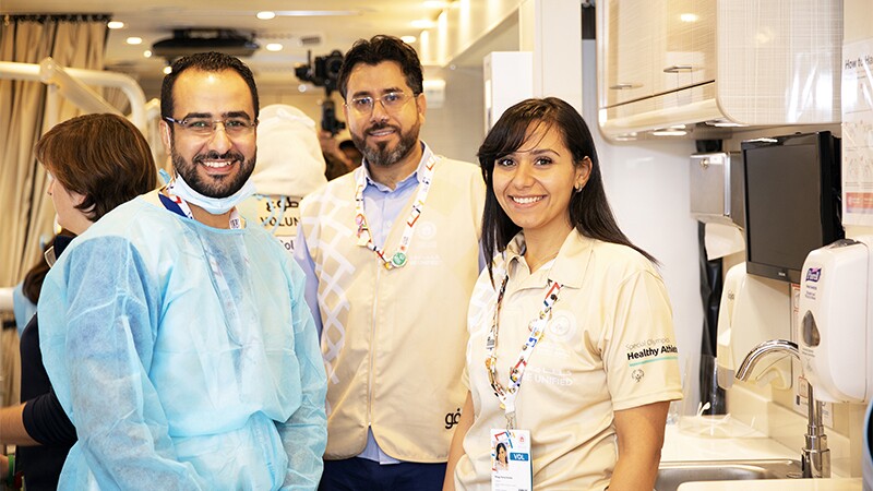 Three volunteers posing for a picture during Healthy Athletes screening, one man in paper scrubs and the other two in tag Healthy Athletes branded polo and vests.