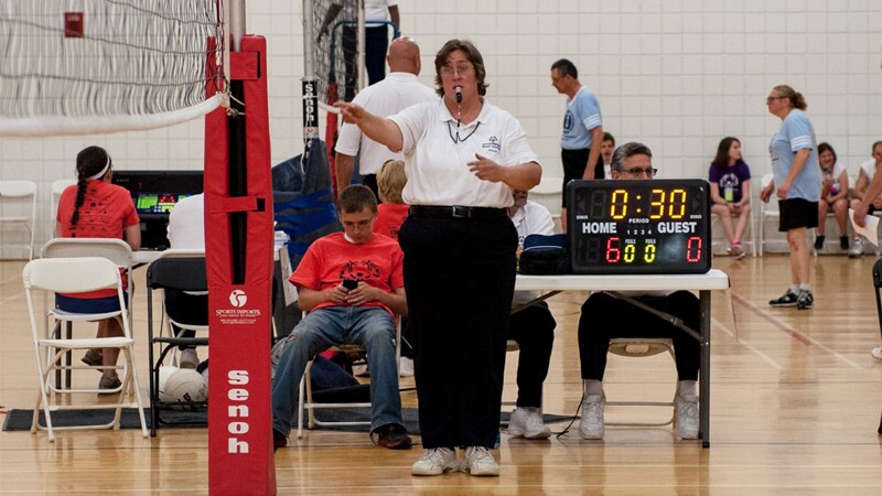 Kristine Hughes officiating a volleyball game. She's standing on the court at one end of the net with her whistle in her mouth.