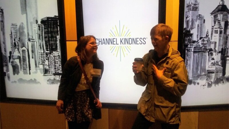 Hanna Joy Atkinson standing with a young man in front of a company logo that reads: Channel Kindness.