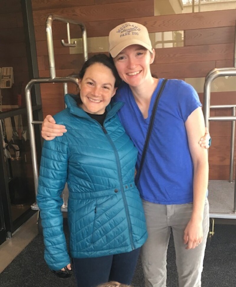 Two women stand with their arms around each other and smile for the camera.