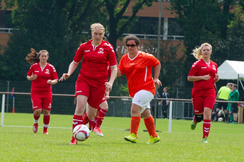 Four women - three in red kits and one in orange and while - running after a football/soccer ball on a football pitch.