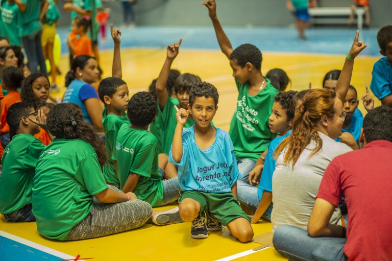 A boy sits in the middle of a group of children and adults, raising their hands and looking directly into the camera, smiling. 