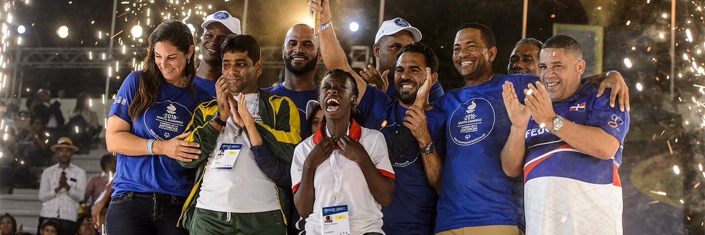 A group of athletes and representative celebrate as fireworks go off in the background.