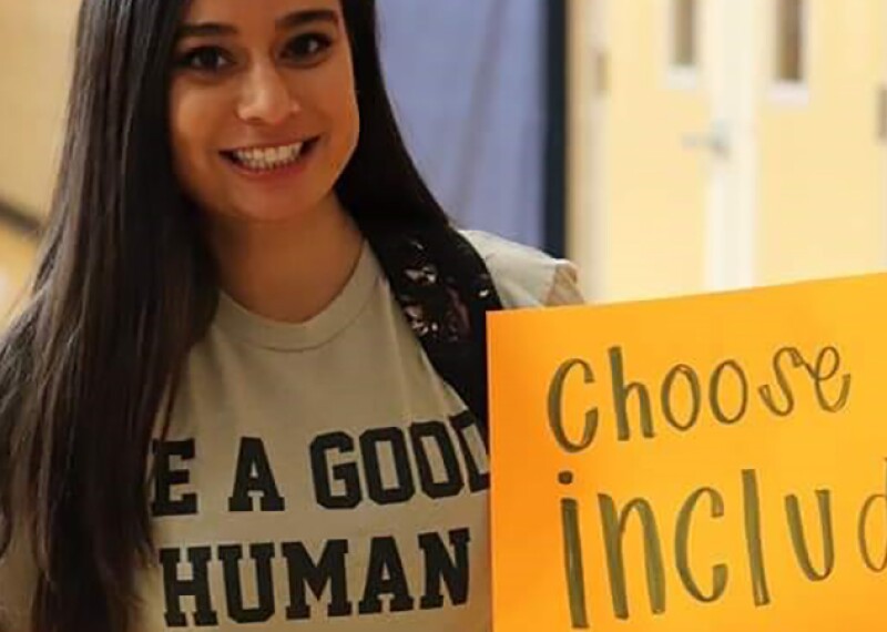 Savyna smiling wearing a shirt that reads, "Be a good human" and holding a sign that reads, "Choose to Include." 