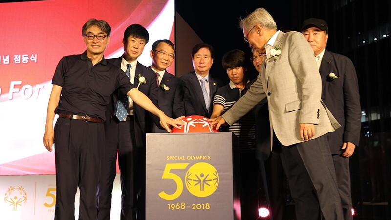 Special Olympics East Asia representatives on stage behind a podium with the Special Olympics 50th Anniversary logo on the front, have their hands on a red Special Olympics Unified Sports decoration. In the background is a red lit up screen.