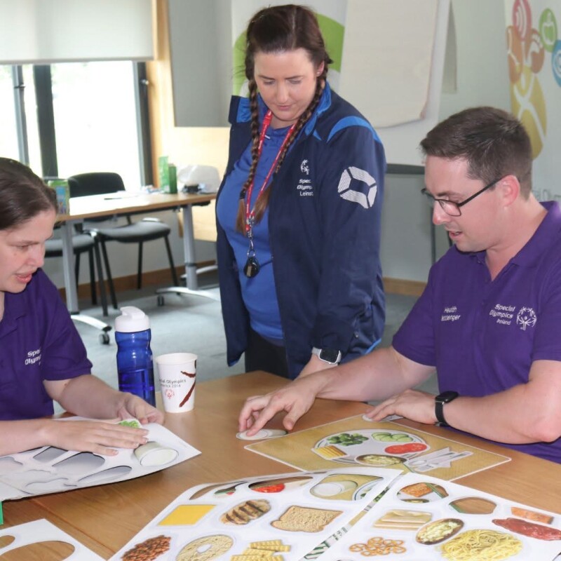 Three individuals around a table, two sitting down and one standing, reviewing photos of different types of foods.
