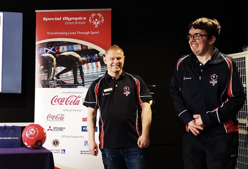 Two Special Olympics Great Britain athletes smiling on a stage standing beside a red Special Olympics Unified ball and a Special Olympics branded pull-up banner.png