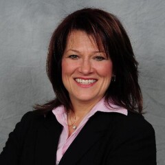 Sharon Bollenbach smiling for a professional photo in a black suite and pink shirt in front of a gray sky background.