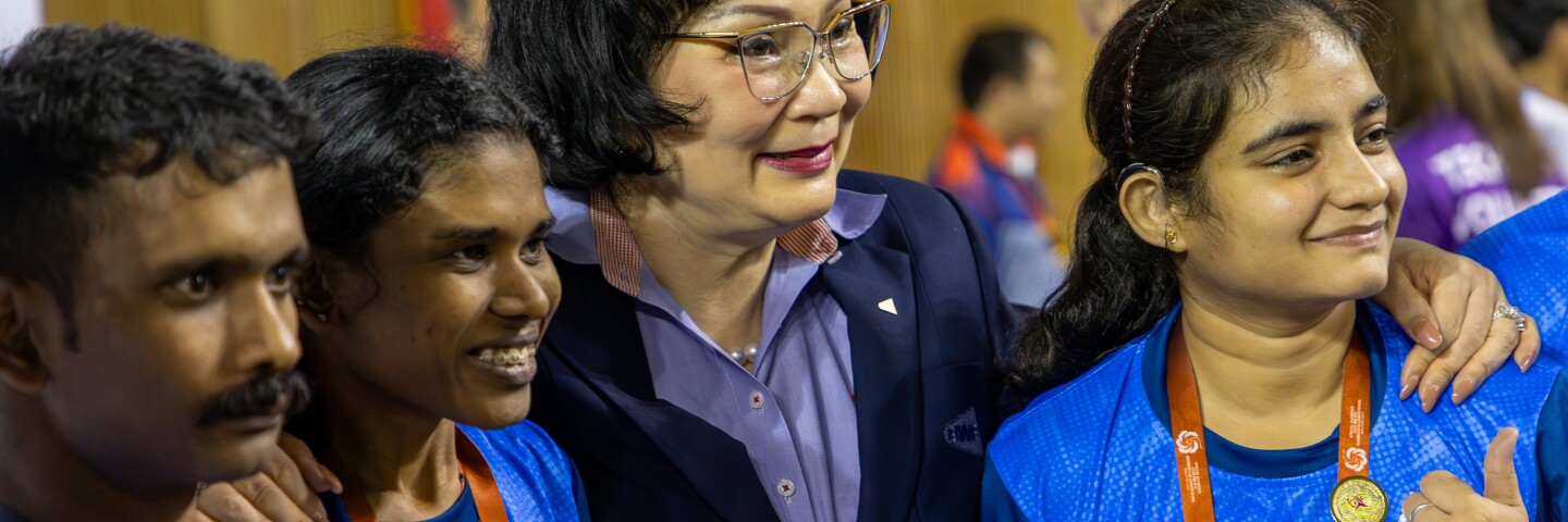Three athletes in blue India jerseys smile and pose with BWF President Khunying Patama Leeswadtrakul.