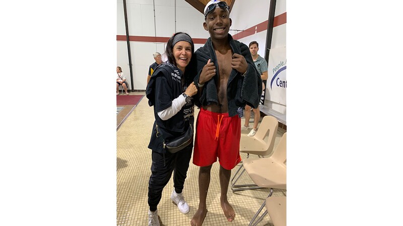Young man (Nick) stands next to a woman on the side of the pool; he has a towel wrapped around him. 