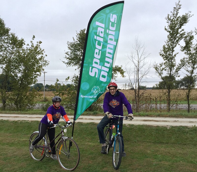 Daniel Smrokowski with friend Georgis in helmets and purple event shirts sitting on bicycles in front of a Special Olympics Illinois marker flag.