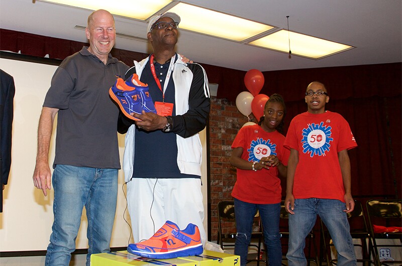 George Piece (on the left) and Bob Beamon (on the right) speak at the Southside Occupational Academy High School in Chicago. Two athletes are in the background watching George and Bob present the shoes to an audience.