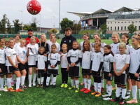 A group of women and girls in football kit stand together on a green pitch while a red football bounces in the air above them.