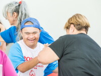Two participants dance together during a Special Olympics Dance Olympics event. One participant wearing a Special Olympics shirt and blue cap holds hands with another participant as they take part in an inclusive dance activity, while others dance nearby.