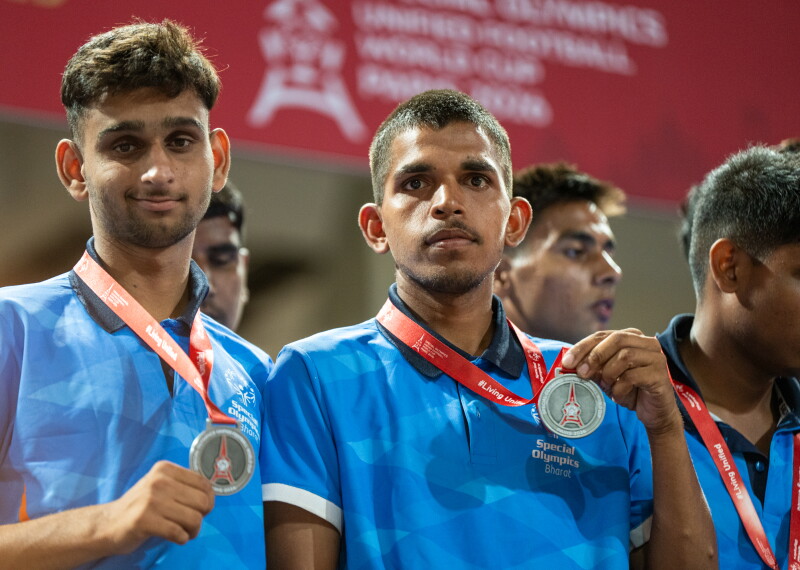 Two athletes in blue team jerseys hold up silver medals while standing together during a medal ceremony.