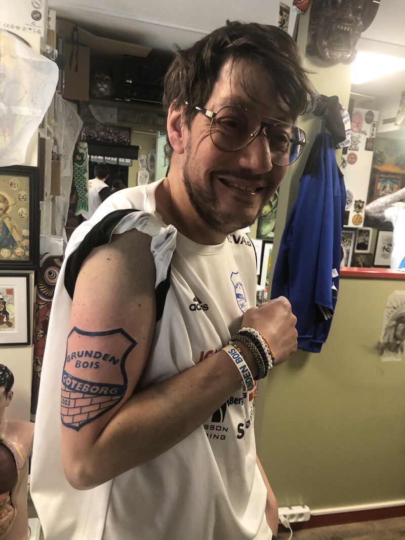 A man with glasses and a white soccer shirt smiles as he shows off a dark blue Grunden Bois tattoo on his upper right arm.