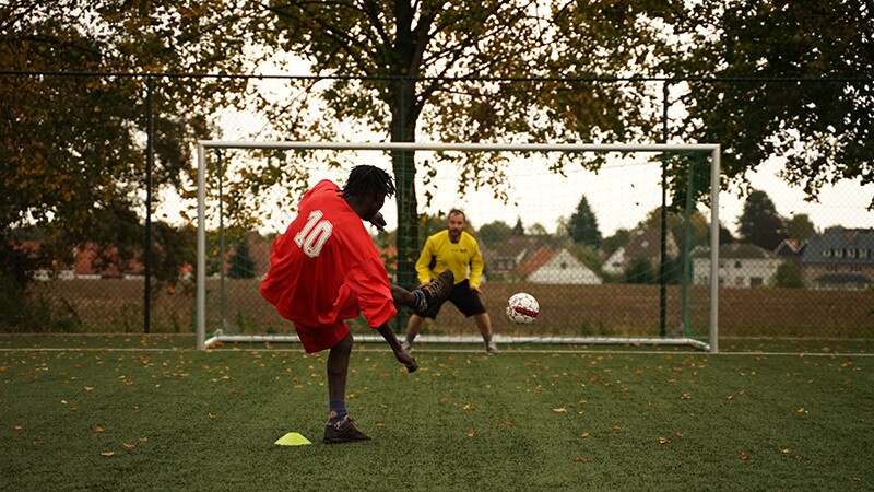 A man in a red football kit with his back to the camera kicks a football towards the net and a goalkeeper in yellow.