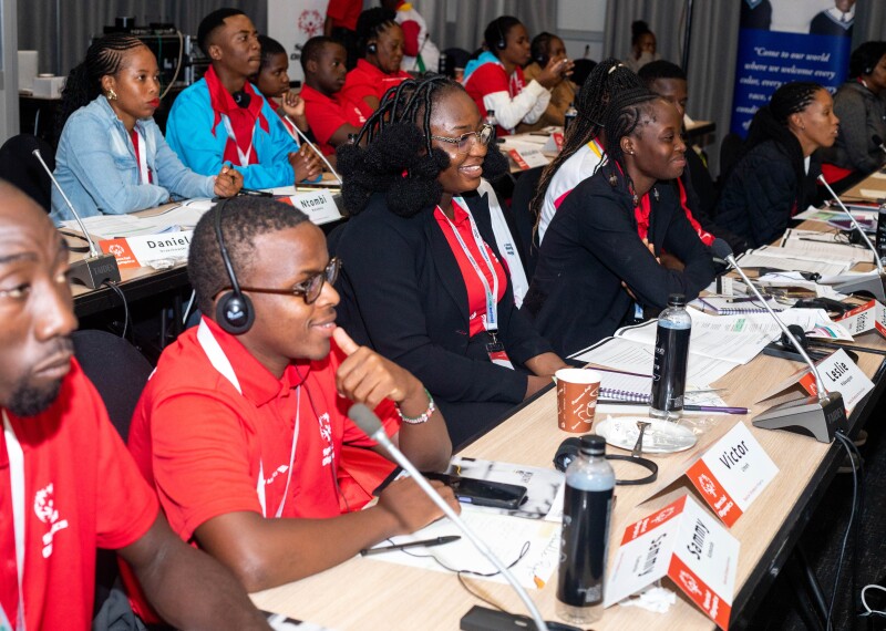 Youth leaders sit along two summit rows. Their desks have microphones and name tags. They are smiling.