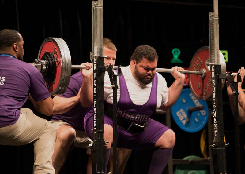 male lifter performs a squat at the rack while coaches and officials assist.
