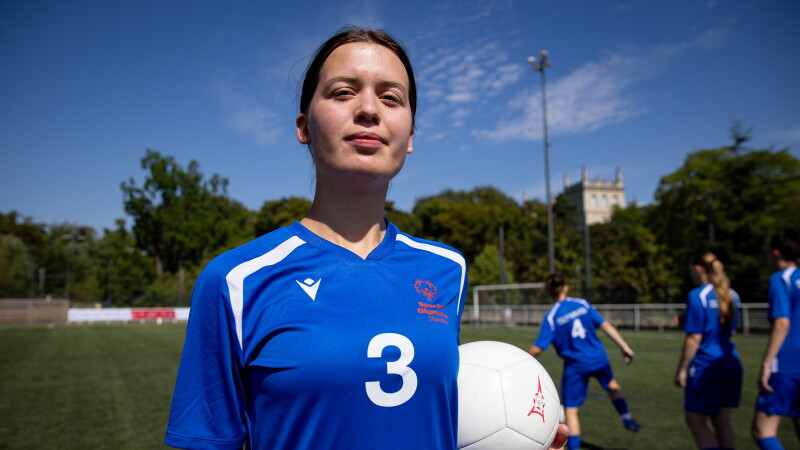 A player wearing a blue Slovakia jersey with the number 3 holds a white football while standing on an outdoor pitch. Teammates practice in the background under a clear blue sky.