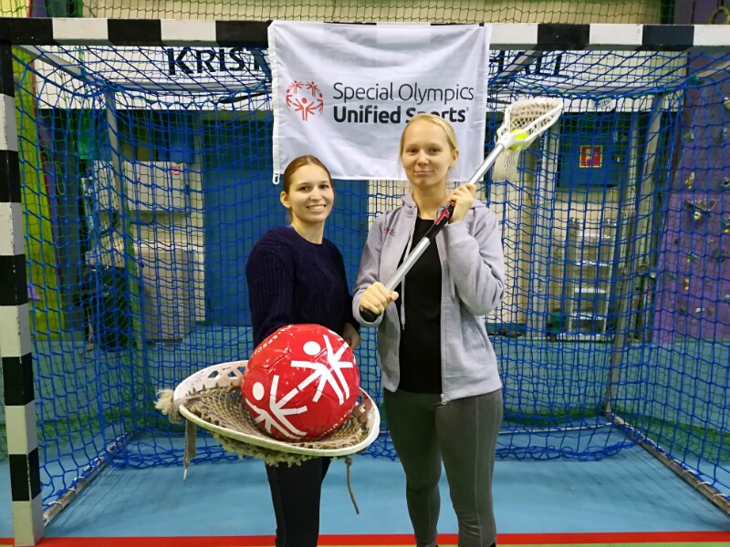Two girls with Lacrosse stick and ball standing in front of net.