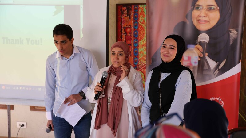 Woman standing with two people on stage in front of signage giving a speech. 