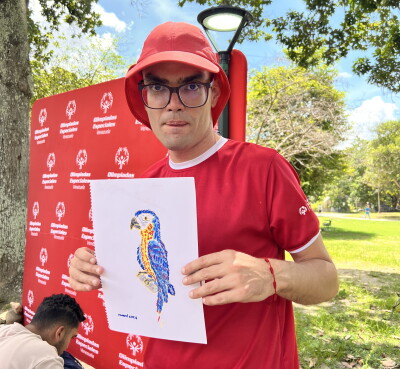 A participant wearing a red Special Olympics Venezuela shirt and hat stands outdoors in a park, holding a drawing of a colorful parrot. A Special Olympics Venezuela backdrop is displayed behind the participant, with trees, grass, and event attendees visible in the background.