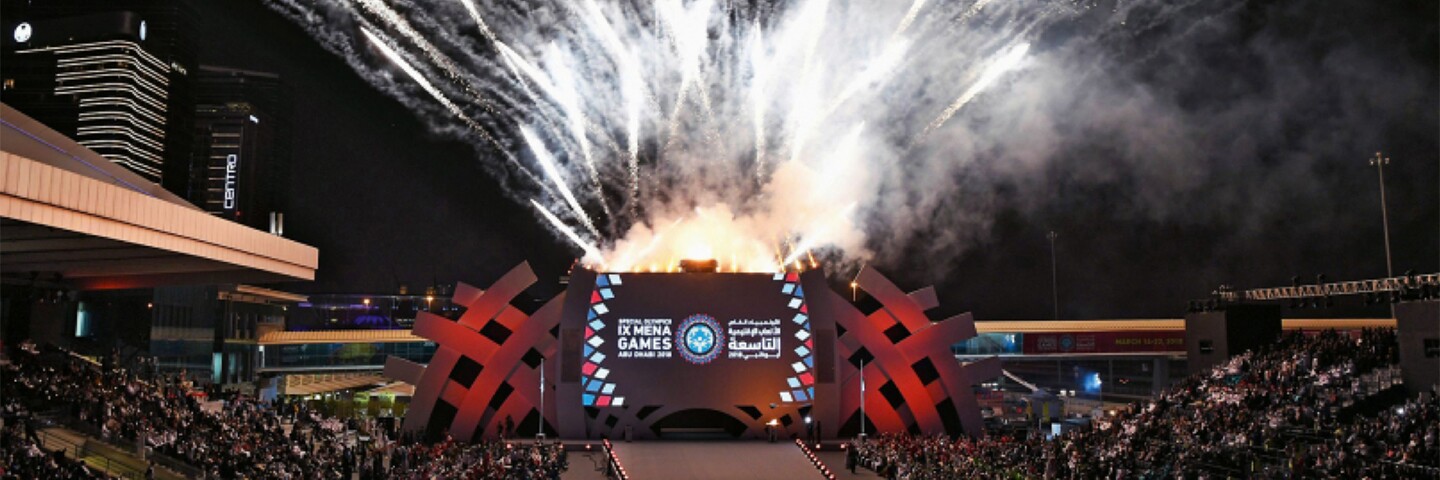 A crowd is watching as fireworks going off behind a stage with a backdrop that reads: IX MENA Games.