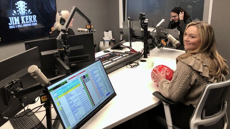 Photo of Elizabeth Rhöm sitting in a chair and holding Special Olympics soccer ball, with Jim Kerr and Shelli Sonstein in the background at Q104.3 FM’s radio station recording room.